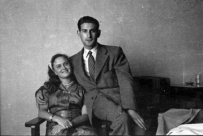 A black and white studio-style portrait of a man and a woman. The man wears a dark suit and tie, while the woman wears a dark...