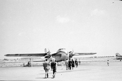 A twin-engine propeller airplane is parked on a tarmac. A group of people, mostly men in suits or light-colored clothing, are...