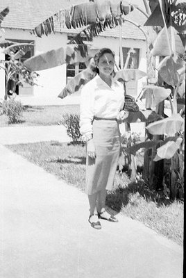 A black and white photograph of a woman standing on a light-colored path. She is wearing a light-colored long-sleeved blouse,...