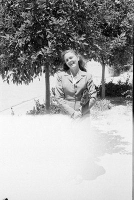 A black and white photograph of a woman smiling, wearing a light-colored button-up blazer and holding a small clutch bag. She...