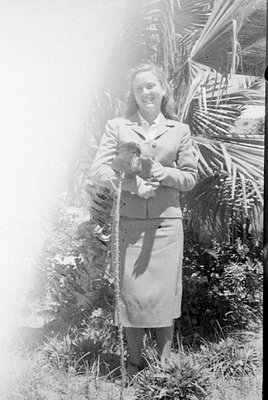 A black and white photograph of a woman standing outdoors among tropical foliage, including palm fronds. She is wearing a lig...