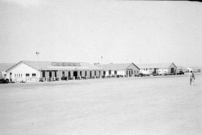 A wide shot of a row of single-story white buildings with gabled roofs under a pale sky. A person walks across a large, flat,...
