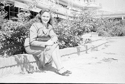 A black and white photograph of a woman sitting on a low concrete ledge. She is wearing a light-colored suit jacket, a matchi...