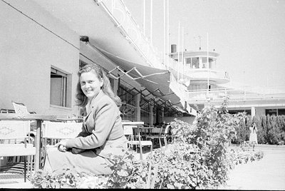 A black and white photograph oriented vertically, showing a person leaning out of a window. The scene includes a balcony with...