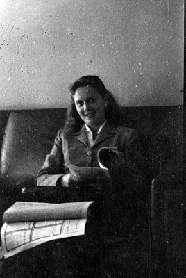 A black and white photograph of a woman sitting on a dark leather sofa. She is wearing a light-colored blazer over a collared...