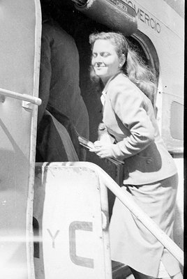 A woman in a light-colored suit and skirt stands in the doorway of a vehicle, holding papers. The word 'MERCIO' is partially ...