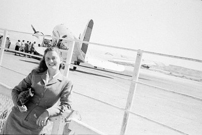 A woman in a tailored suit and skirt smiles while leaning against a white railing. In the background, a propeller airplane is...