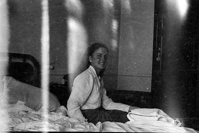 A black and white photograph of a woman sitting on a bed with white linens. She is wearing a light-colored long-sleeved shirt...