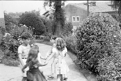 Four children stand in a circle holding hands on a paved path. They are surrounded by large bushes and flowers, with a two-st...