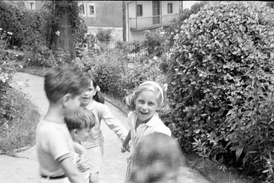 Three children stand on a paved garden path. A boy in a light-colored t-shirt is in the foreground, while a girl with a headb...