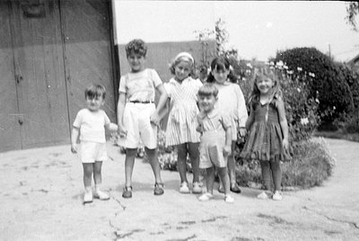 Five children stand in a row on a paved outdoor surface. They are dressed in mid-century style clothing, including shorts, a ...