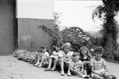 Six children sit in a row on a paved surface outdoors. They are positioned in front of a dark wall and flowering bushes. The ...