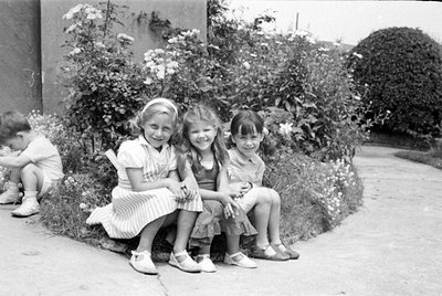 Three young girls sit side-by-side on a garden border in front of flowering bushes. A fourth child is partially visible on th...