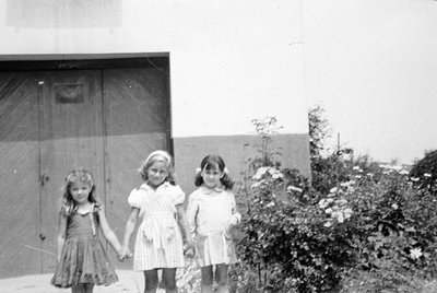 Three young girls stand side-by-side in front of a light-colored building with large wooden doors. They are wearing light-col...