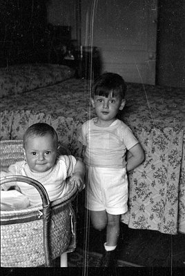 A black and white photograph of two young children in a bedroom. An infant sits in a wicker bassinet in the foreground, while...