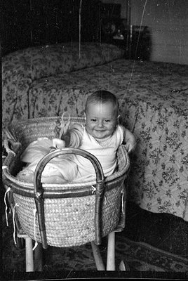 A black and white photograph of an infant sitting in a woven wicker bassinet. The baby is wearing a light-colored garment and...
