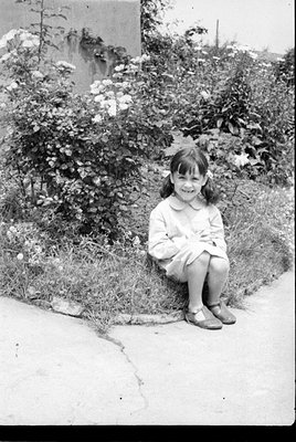 A black and white photograph of a young girl with pigtails sitting on a grassy verge. She is wearing a light-colored dress an...