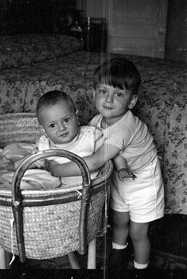 A black and white photograph of a young child standing next to a baby in a woven bassinet. The child has an arm draped over t...