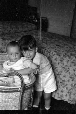 A black and white photograph of two young children. An infant sits in a woven wicker bassinet while an older child in a light...