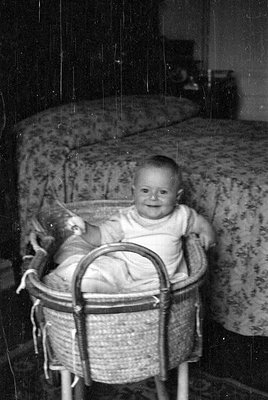 A black and white photograph of an infant sitting in a woven wicker bassinet. The baby is wearing a light-colored one-piece g...