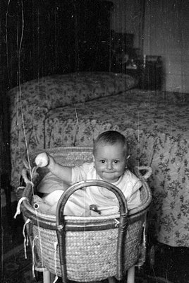 A black and white photograph of an infant sitting in a woven wicker bassinet. The child is holding a small, round object in t...