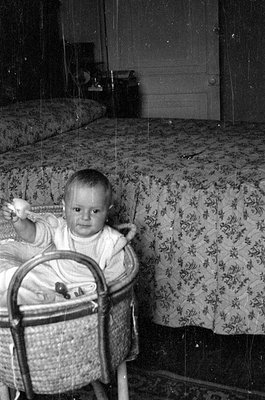 A black and white photograph of an infant sitting in a woven bassinet. The child wears a light-colored garment and holds a sm...