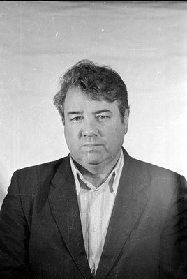 Black and white headshot of a man with wavy hair, wearing a dark suit jacket over a light-colored striped collared shirt. He ...