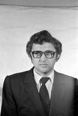 Black and white studio portrait of a man wearing thick-rimmed glasses, a dark suit, and a patterned tie. He is positioned aga...