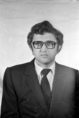 Black and white studio portrait of a man wearing thick-rimmed glasses, a dark suit, a white shirt, and a patterned tie. He ha...