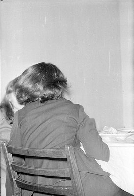 A black and white photograph taken from behind a person with short, wavy hair sitting in a wooden ladder-back chair. The pers...