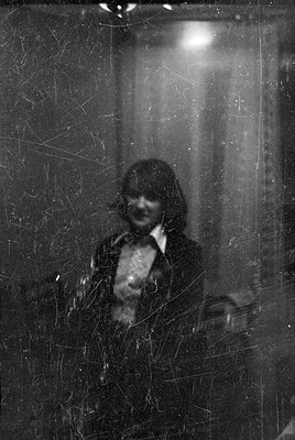 A black and white portrait of a person with dark, shoulder-length hair wearing a dark jacket over a light-colored collared sh...