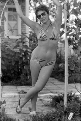 A black and white photograph of a woman wearing a striped bikini and sunglasses. She is posing with her arms raised, holding ...
