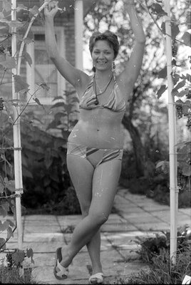 A black and white photograph of a woman in a striped two-piece swimsuit and sandals. She is reclining outdoors under a trelli...