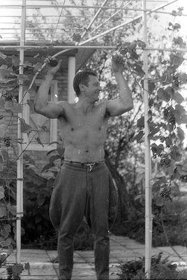 A shirtless man stands under a metal trellis with climbing vines. He is wearing trousers and has both arms raised, holding on...