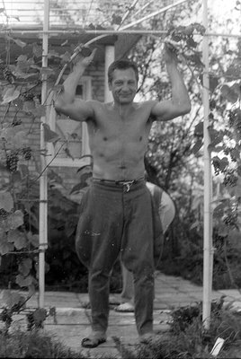 A shirtless man stands outdoors under a white metal trellis entwined with grapevines. He is wearing high-waisted trousers and...