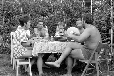 A black and white photograph of four people sitting around a table covered with a patterned cloth in an outdoor garden settin...