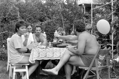 A group of six people sit around a table covered with a lace tablecloth in an outdoor garden setting. A white balloon floats ...
