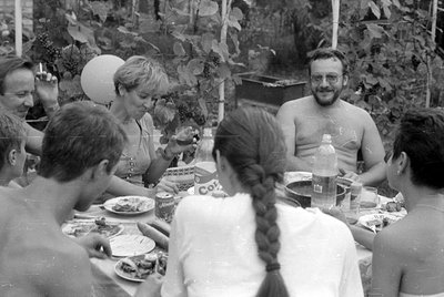 A group of people gathered around a table outdoors with food and drinks. A man and woman are shirtless, and a woman with a lo...