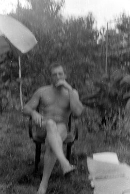 A shirtless man sits in a chair outdoors, legs crossed, with his hand near his mouth. A light-colored umbrella is visible in ...