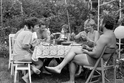 A group of people sit around a table covered with a lace tablecloth in an outdoor garden setting. Several individuals are shi...