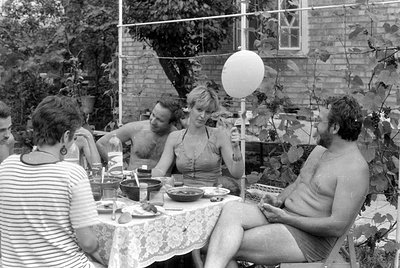 A group of people are gathered around a table covered with a lace tablecloth in an outdoor garden setting. A shirtless man si...