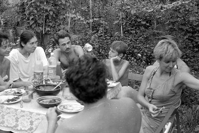 A group of people sit around a table covered with a lace cloth in an outdoor garden setting. Bowls of food and plastic bottle...