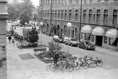 A street scene featuring a stone building with storefronts labeled 'ORION FUR'. Several cars are parked along the curb, and a...