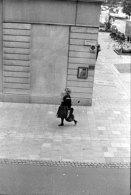 A black and white street photograph of a person walking past a stone building facade. The person is carrying two large dark b...