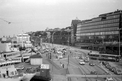 A waterfront road with heavy traffic, including buses and cars, runs alongside a large glass-fronted modern building. To the ...