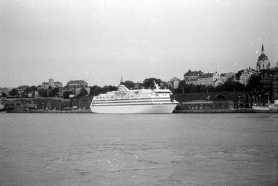 A large white cruise ship is docked at a waterfront. In the background, a cityscape features several multi-story buildings an...