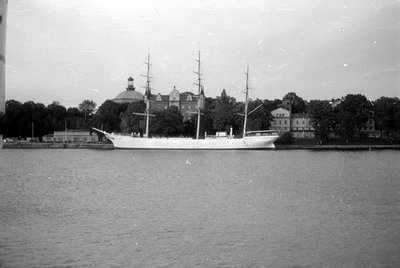 A large white three-masted sailing ship is docked along a waterfront. In the background, a large building with a prominent do...