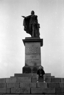 A black and white photograph of a large stone monument topped with a statue of a man in a cloak. A man in a dark jacket and l...