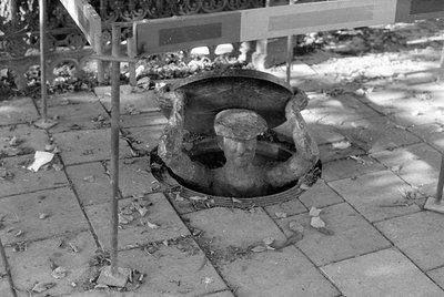 A black and white photograph of a sculpture depicting a man in a cap emerging from a manhole in a paved ground. The figure is...