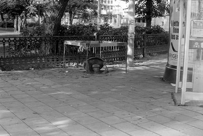 A black and white photograph of a city sidewalk. A metal table stands over a circular manhole cover, next to a decorative iro...